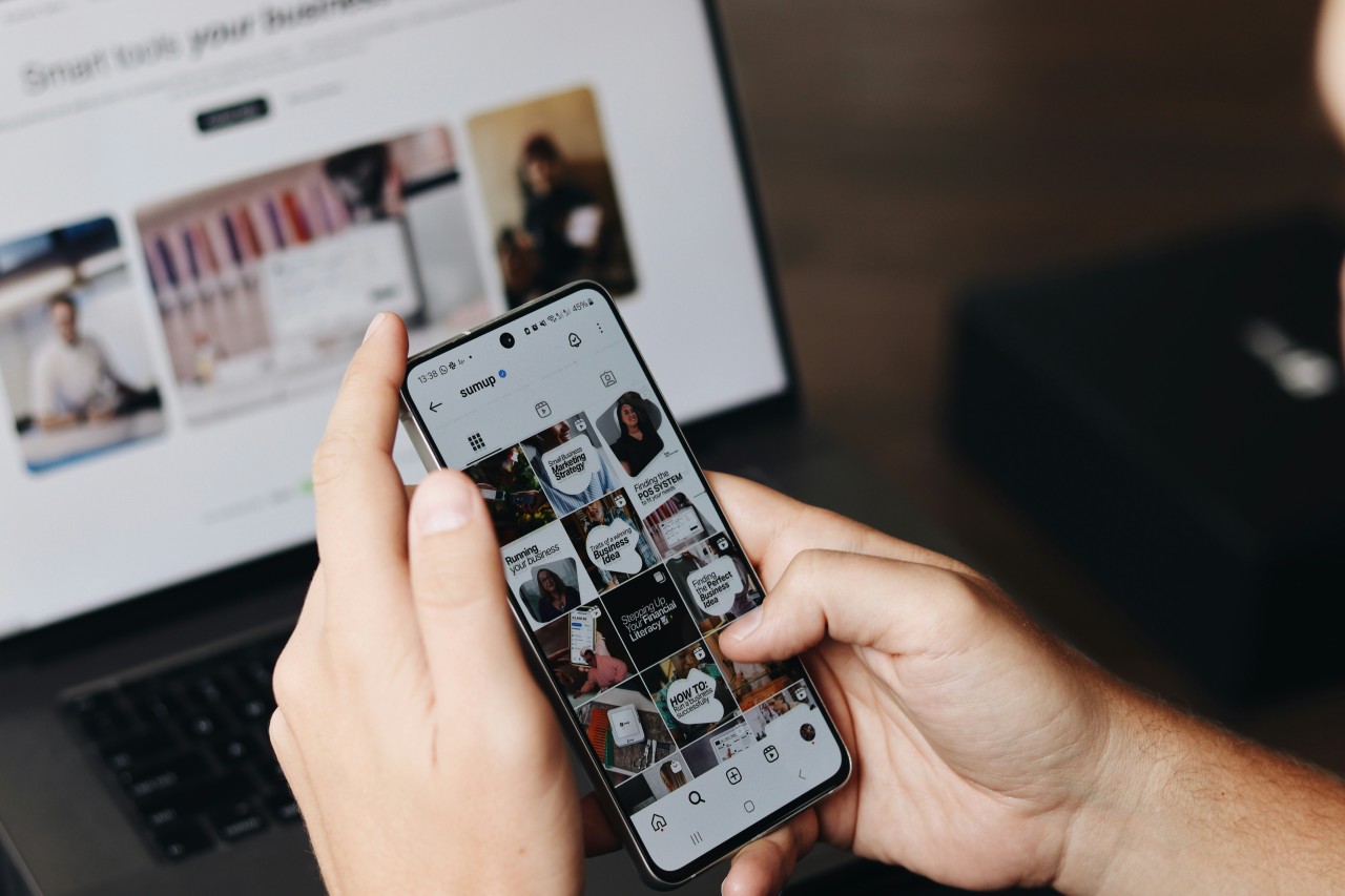 Person holding a smartphone displaying the SumUp Instagram profile grid, with a laptop showing the SumUp website open in the background.